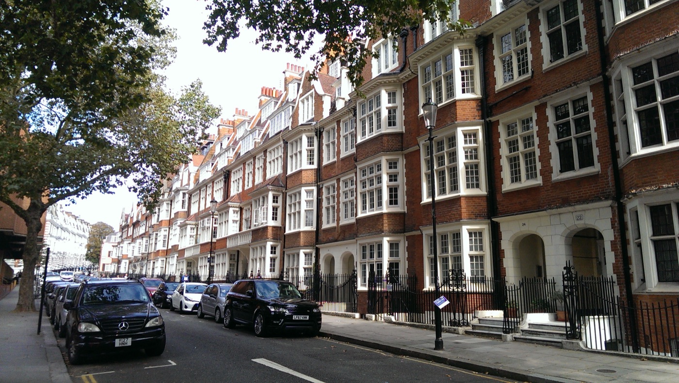 A terrace of red brick mansion flats with white bay windows on a tree-lined London street